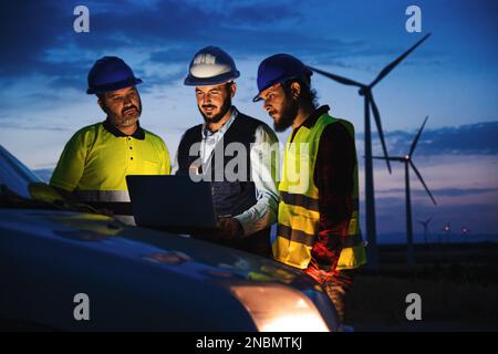 Team work of three renewable energy engineers with helmets and protection equipment, working and planing at night with laptop computer on top of the car with a wind power generators in the background. Renewable energy concept. High quality photo Stock Photo