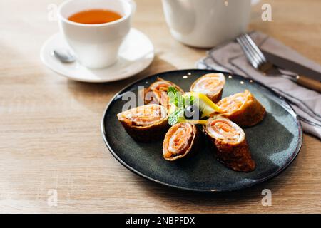 Rolls of thin pancakes with smoked salmon and cream cheese on dark plate on wooden table. Delicious and healthy breakfast or brunch Stock Photo