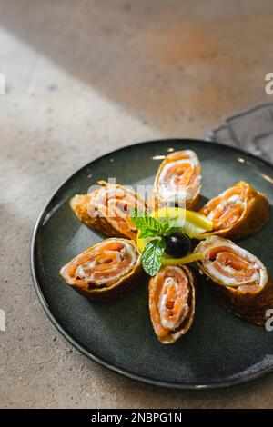 Rolls of thin pancakes with smoked salmon and cream cheese on dark plate on table. Delicious and healthy breakfast or brunch. Copy space Stock Photo