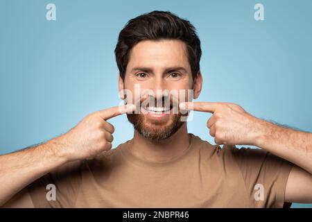 Handsome bearded man in t-shirt posing over blue background, smiling and pointing with fingers at perfect teeth Stock Photo