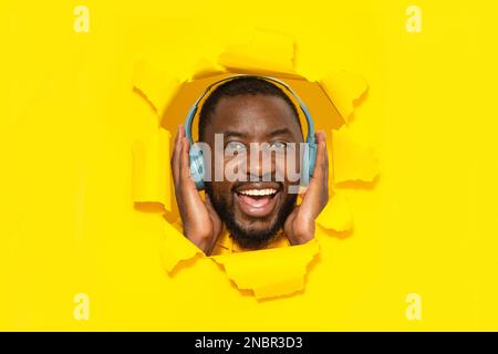 Excited black man touching wireless headphones and listening to music online, smiling through hole in torn yellow paper Stock Photo