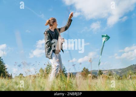 Smiling teenager boy with flying colorful kite on the high grass meadow in the mountain fields. Happy childhood moments or outdoor time spending conce Stock Photo
