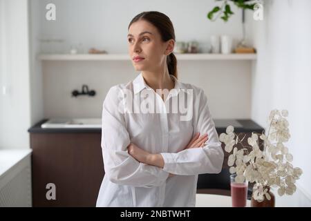 Close-up of young beautiful smart business caucasian brunette woman wearing business style white shirt standing with folded arms in a bright room. Stock Photo