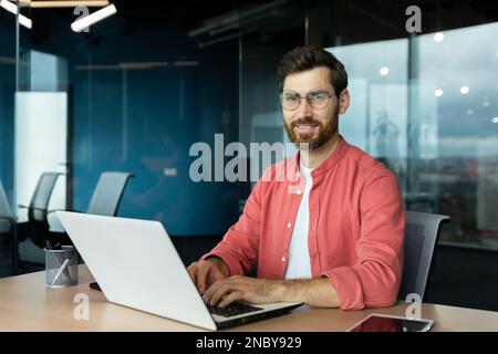Close-up portrait of mature businessman inside office, man with beard and red shirt smiling and looking at camera, programmer developer coding software at workplace with laptop. Stock Photo