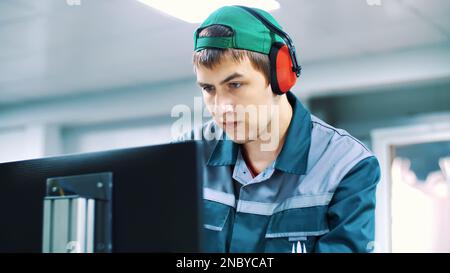 portrait of an employee of an enterprise, factory. a worker in red headphones from noise, looks at a computer monitor, adjusts, controls work of machines, automated production. High quality photo Stock Photo