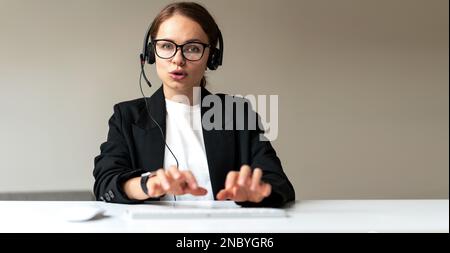 Female consultant of call center in headset speaking into headset microphone and typing comments on keyboard. Stock Photo
