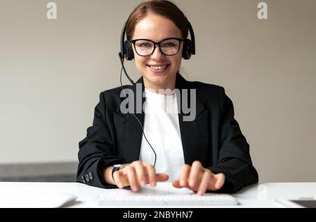 Worker customer service online center woman in headset smiling typing comments on keyboard while talking with customer online. Stock Photo