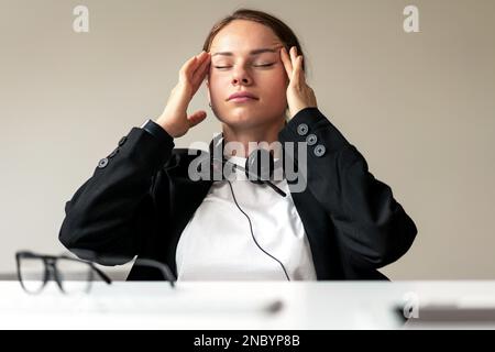 Tired businesswoman sitting at desk with eye closed rubbing temple area with her hands. Stock Photo