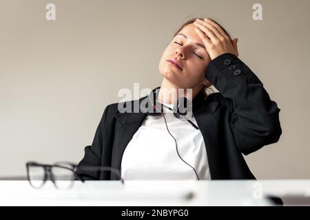 Woman office worker sitting at desk has headache touching forehead with her hand, feeling unwell at work. Stock Photo