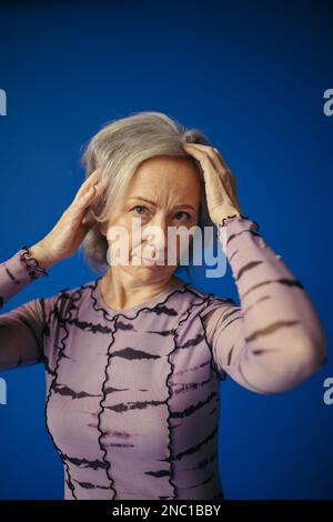 portrait of senior woman in purple dress adjusting grey hair while posing isolated on blue Stock Photo