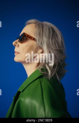 profile of senior grey haired lady in trendy sunglasses and green leather jacket isolated on blue Stock Photo