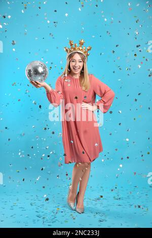 Happy young woman in party crown with disco ball and confetti on light blue background Stock Photo