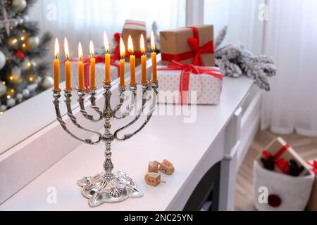 Silver menorah near dreidels with symbols He, Pe, Nun, Gimel on white mantelpiece in room decorated for Christmas Stock Photo