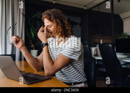 Young curly man touching his nose bridge while using laptop by desk in office Stock Photo