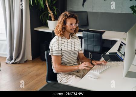 Young employee working on desktop computer and laptop while sitting at table in modern office Stock Photo