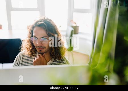 Young curly man with eyeglasses using laptop while sitting by desk in office Stock Photo