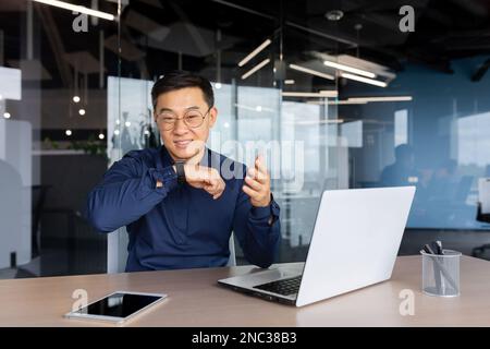 A young Asian businessman, a freelancer works in the office using a laptop. He talks through the smart watch on his arm. Stock Photo