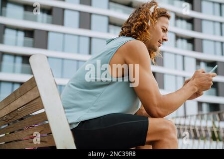 Young handsome focused sporty long-haired man in headphones with phone sitting on the bench outdoors Stock Photo