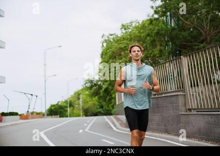 Young handsome athletic long-haired man in headphones running outdoors Stock Photo