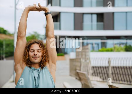 Young athletic long-haired handsome smiling man with closed eyes stretching his arms , while standing outdoors Stock Photo