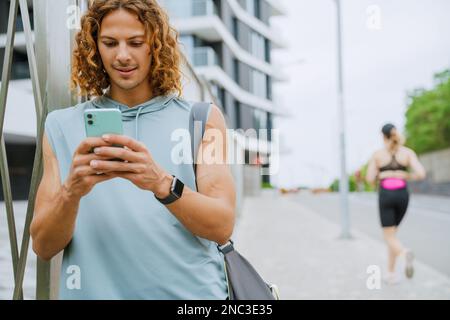 Portrait of young long-haired smiling athletic man with bag using phone , while standing outdoors Stock Photo