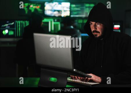 Hooded male criminal hacker using a laptop to breack the security of the firewall, at background a lot of computer screens. Pishing concept. High quality photo Stock Photo