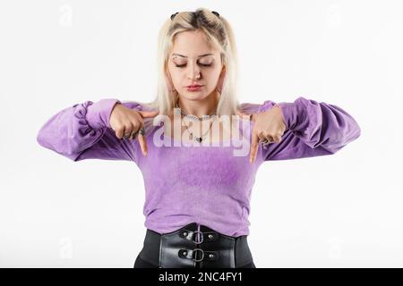 Portrait of young blonde woman wearing ribbed blouse standing isolated over white background pointing downward looks dissatisfied. Pointing downwards Stock Photo