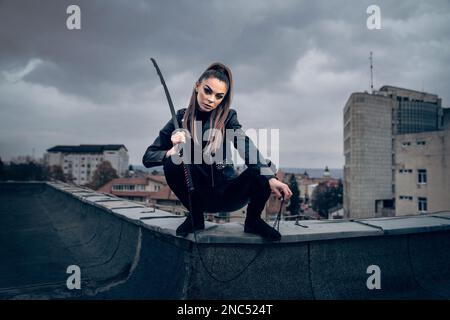 A cool female ninja warrior in leather outfit wielding a Japanese sword on the top of a building Stock Photo