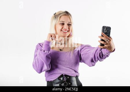 Young blonde girl smiling happy wearing ribbed blouse isolated over white background makes a video call with a smartphone and points at herself with h Stock Photo