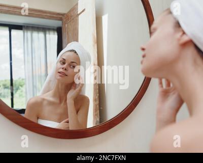 A beauty woman stands in front of a mirror after a shower in a towel on her head looks at her reflection and does a facial massage applies a day cream Stock Photo