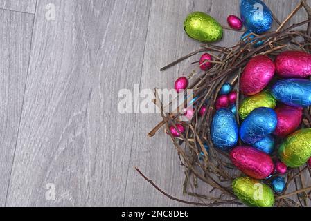 Foil wrapped multi coloured easter eggs in pink, green, blue and yelow in a natural nest made of sticks and twigs, against a grey wooden background. Stock Photo