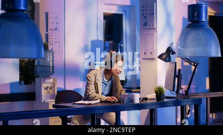 Cheerful employee working on research with headset, listening to music while typing economy data on computer. Young adult planning investment before deadline, wearing headphones. Stock Photo