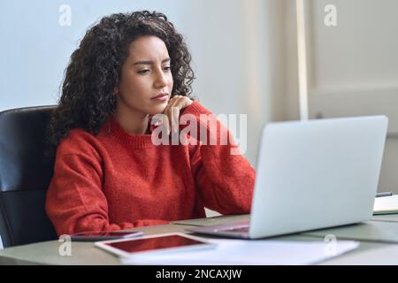 Young latin business woman working on laptop thinking analyzing online data. Stock Photo