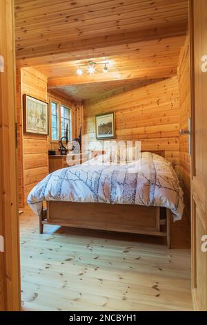 Queen size bed with wood frame and striped bedspread and matching pillows in long and narrow bedroom inside piece sur piece Scots pine log home. Stock Photo