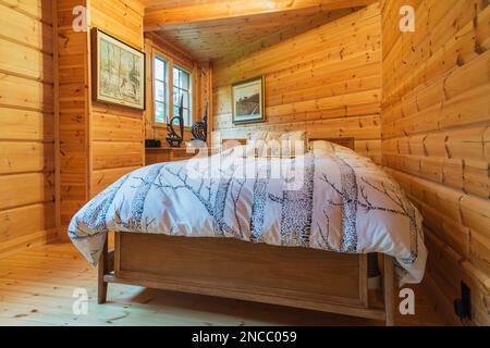 Queen size bed with wood frame and striped bedspread and matching pillows in long and narrow bedroom inside piece sur piece Scots pine log home. Stock Photo