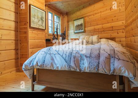 Queen size bed with wood frame and striped bedspread and matching pillows in long and narrow bedroom inside piece sur piece Scots pine log home. Stock Photo