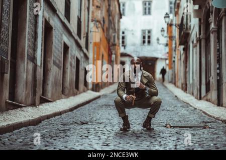 A shot focused on a bald black man dressed in khaki overalls standing in the middle of a black cobblestone street with typical old houses, crouching d Stock Photo