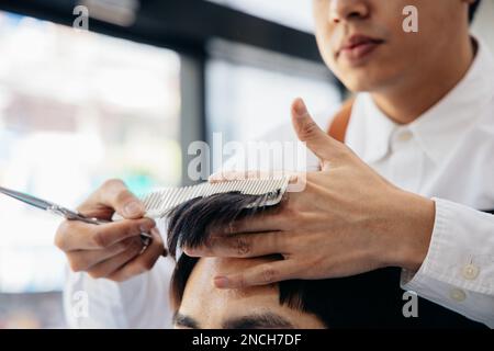 Young and handsome male hairdresser trimming man's hair using comb and scissors while concentrating on work in modern salon Stock Photo