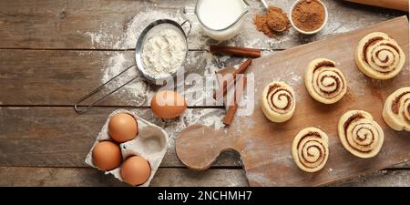 Board with uncooked cinnamon rolls and ingredients on wooden background Stock Photo