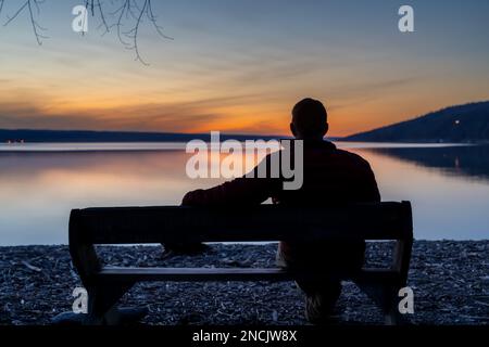 Winter scene of a man with red jacket on a bench looking out over a lake at sunset. Taking time for personal reflection, introspection, thinking about Stock Photo