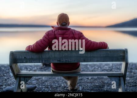 Winter scene of a man with red jacket on a bench looking out over a lake at sunset. Taking time for personal reflection, introspection, thinking about Stock Photo