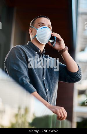 Making business connections in the face of a pandemic. a young businessman wearing a mask and using a smartphone on the balcony of an office. Stock Photo