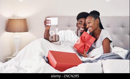 Sharing our special day with our friends online. a young woman holding up a gift box while taking a selfie with her boyfriend. Stock Photo