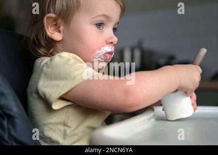 Young Kid Eating Blend Mashed Feed Sitting in High Chair. Baby Weaning. Little Girl Learning to Eat Yogurt, Feeding Himself. Small Hand with Spoon. Br Stock Photo