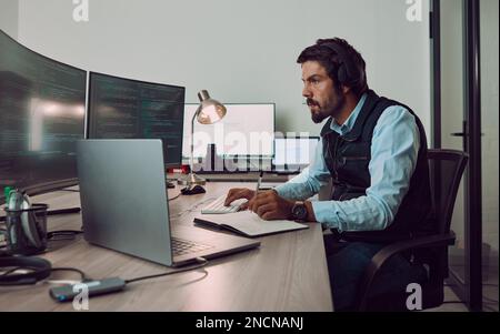 Headphones, programmer and man on computer with book, coding or programming while writing notes at night. Information technology, notebook and male Stock Photo
