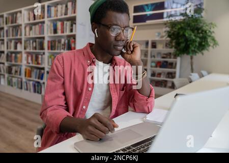 Thoughtful hipster man student or freelancer thinks of online project looking at laptop in library.  Stock Photo