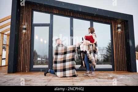 Man in love making marriage proposal to girlfriend under winter snow while woman holding bouquet of flowers. Happy couple sharing romantic moment outside scandinavian house barnhouse. Stock Photo
