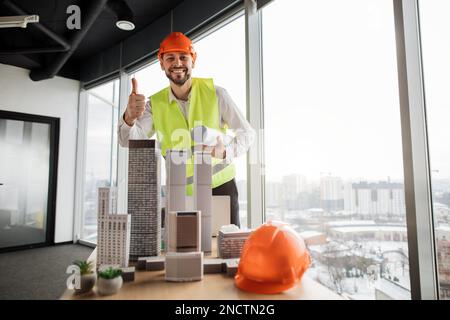 Architect man or male builder adding component to a building model, working on a city district urban planning project showing thumb up at open space office with panoramic windows with blurry cityscape Stock Photo
