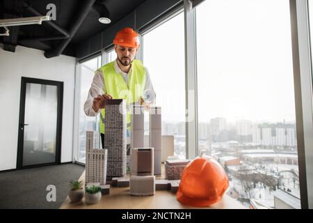 Engineer man in orange helmet and reflective vest standing near table with architecture design of city buildings, residential project maquette. Business man working on common construction project. Stock Photo