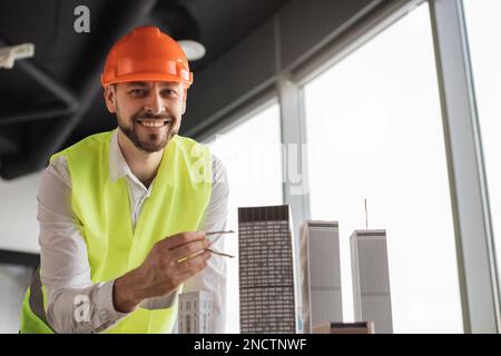 Close up of bearded engineer in protective helmet working on building complex prototype project of residential or business district, measuring city model using compass for drawing at office. Stock Photo